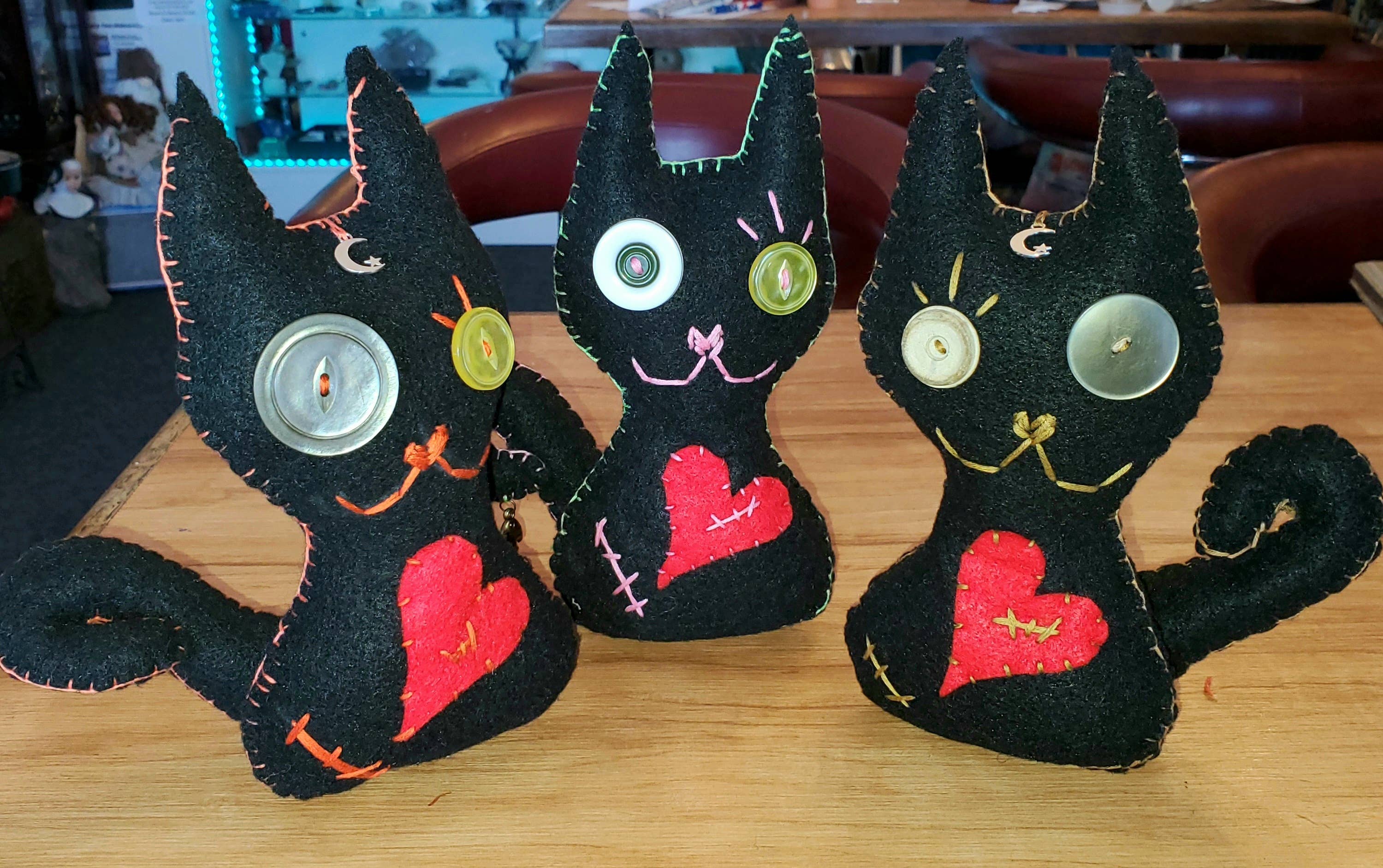 Three handmade black felt cat dolls with large button eyes and red stitched hearts, displayed on a wooden table.