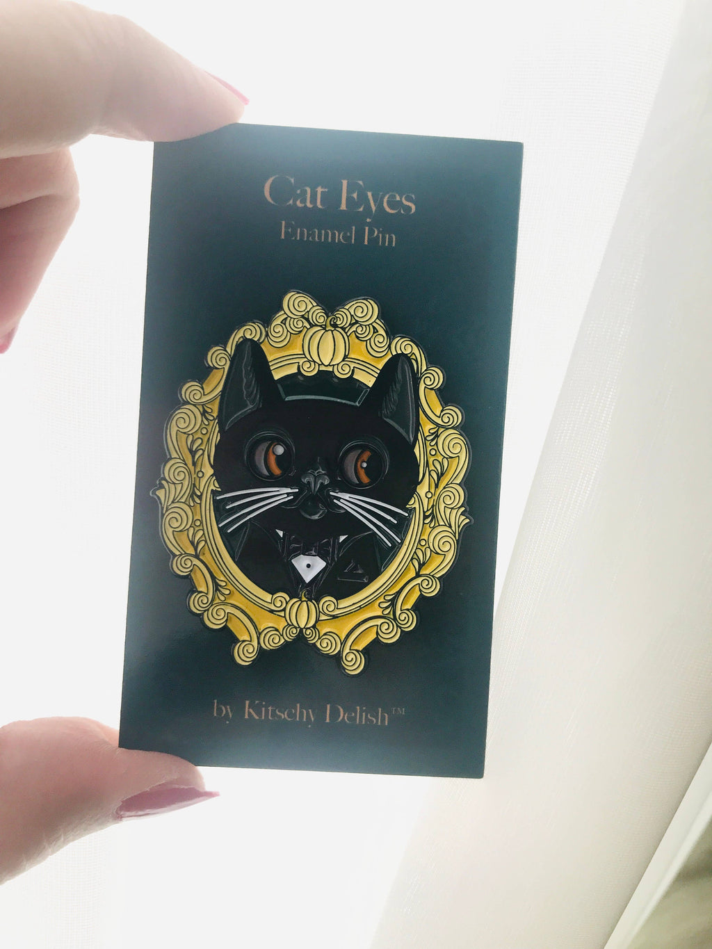 Enamel pin with cat design on a dark green card held by a hand.