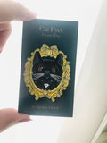 Enamel pin with cat design on a dark green card held by a hand.