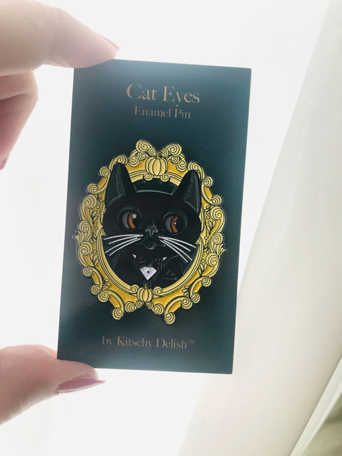 Enamel pin with cat design on a dark green card held by a hand.