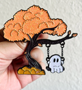 Enamel pin of a tree with a ghost hanging from a branch, held in a hand against a light background. Halloween