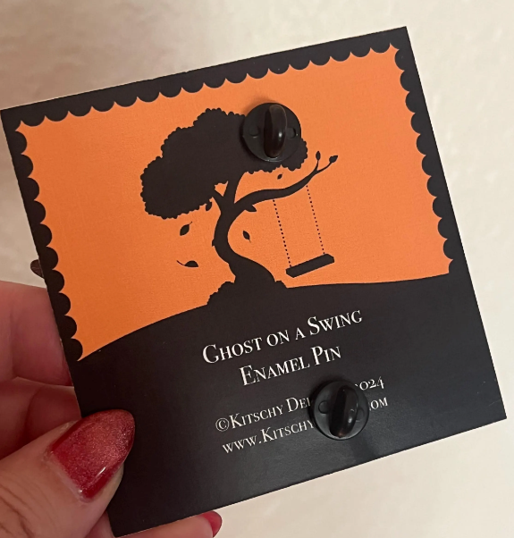 Enamel pin with 'Ghost on a Swing' design on an orange and black card held by a hand.