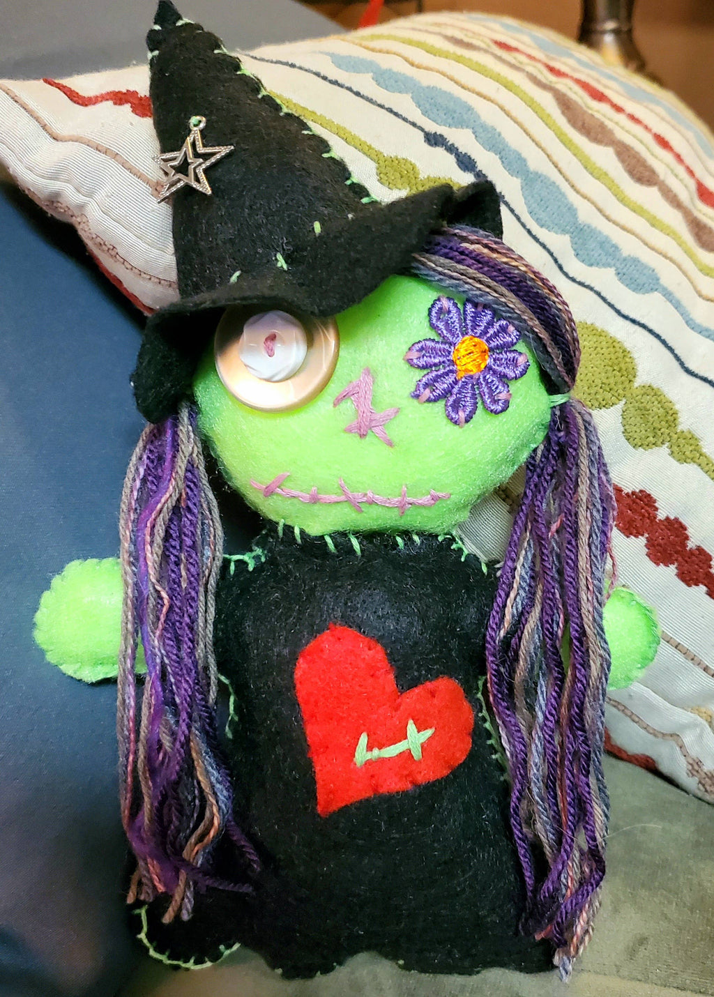 A handmade green felt witch doll with a black pointed hat, purple yarn hair, a red heart on its black dress, and a button eye, resting on a striped pillow.