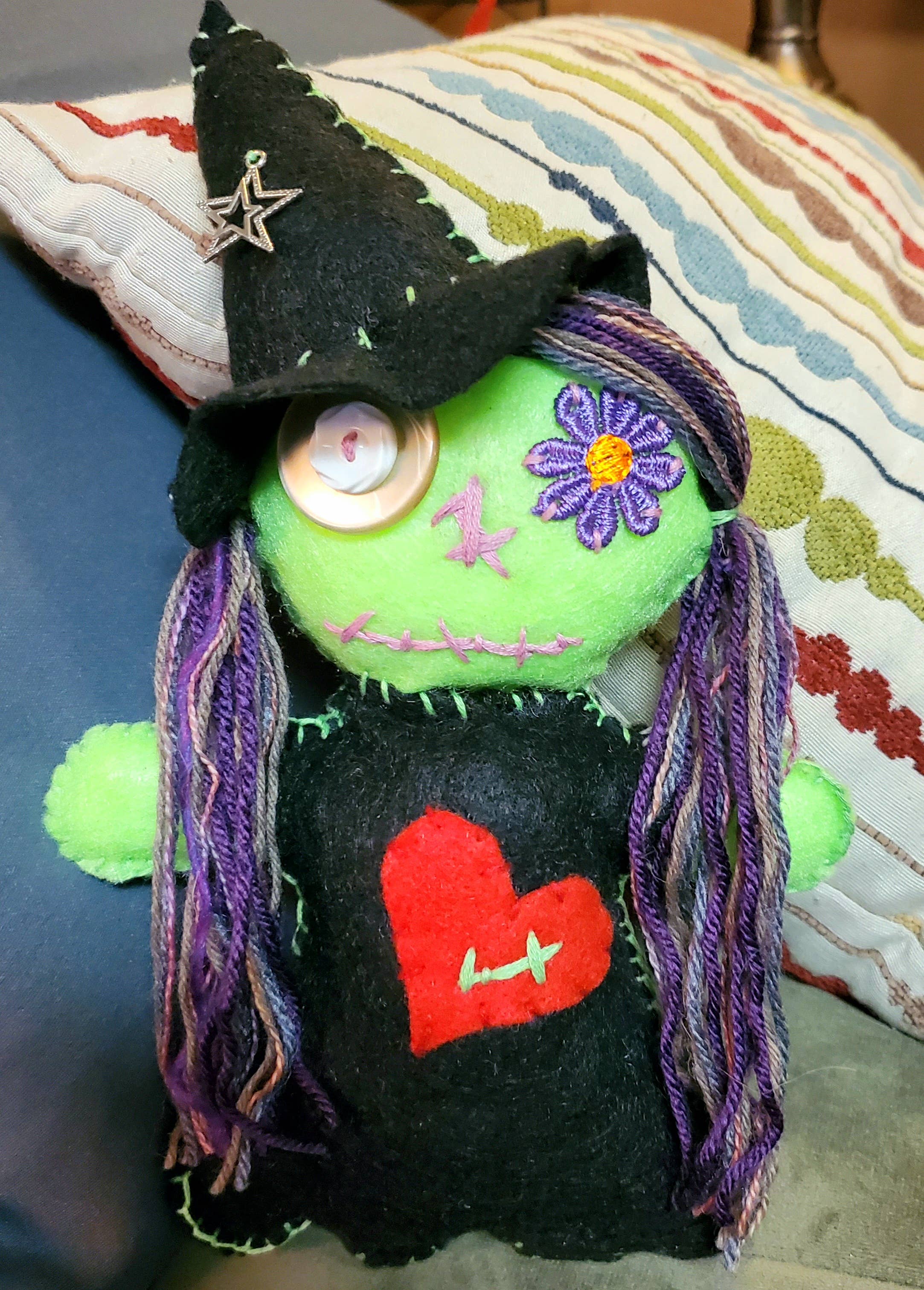 A handmade green felt witch doll with a black pointed hat, purple yarn hair, a red heart on its black dress, and a button eye, resting on a striped pillow.