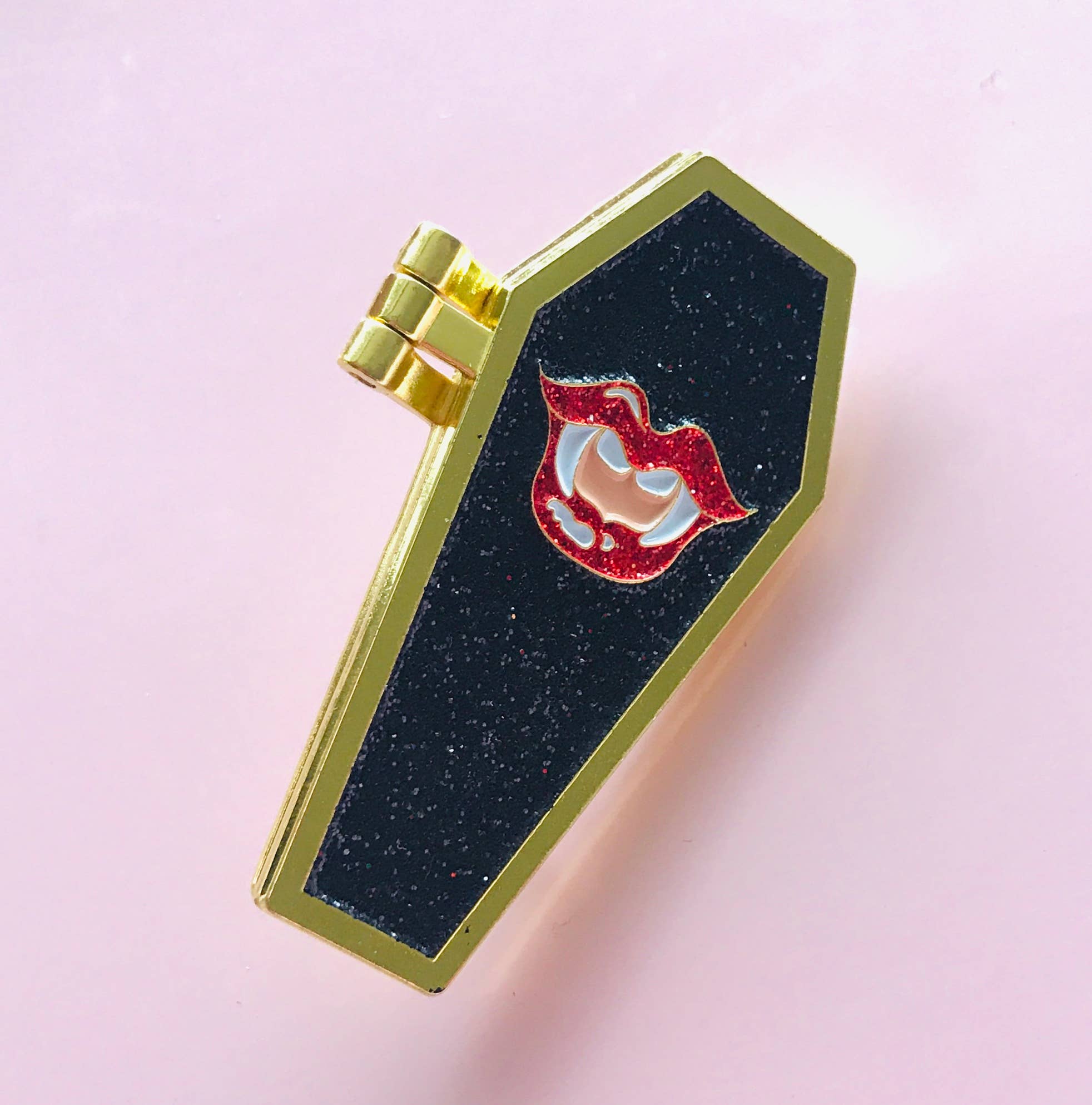 Black coffin-shaped enamel pin with gold trim and red and white design on a pink background