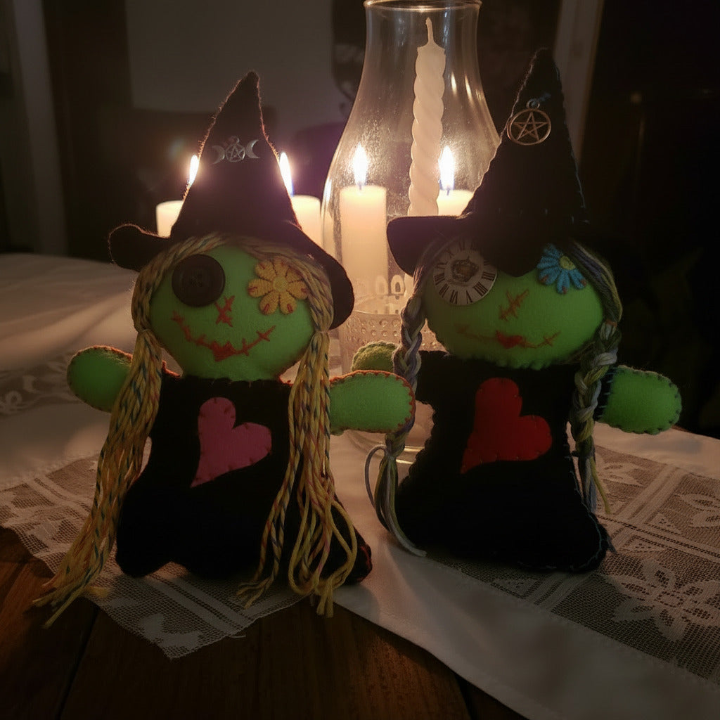 Two handmade witch dolls made of green felt and black fabric, decorated with colorful yarn hair, embroidered faces, and heart-shaped dresses in pink and red, set against a dark background with a glass bottle.