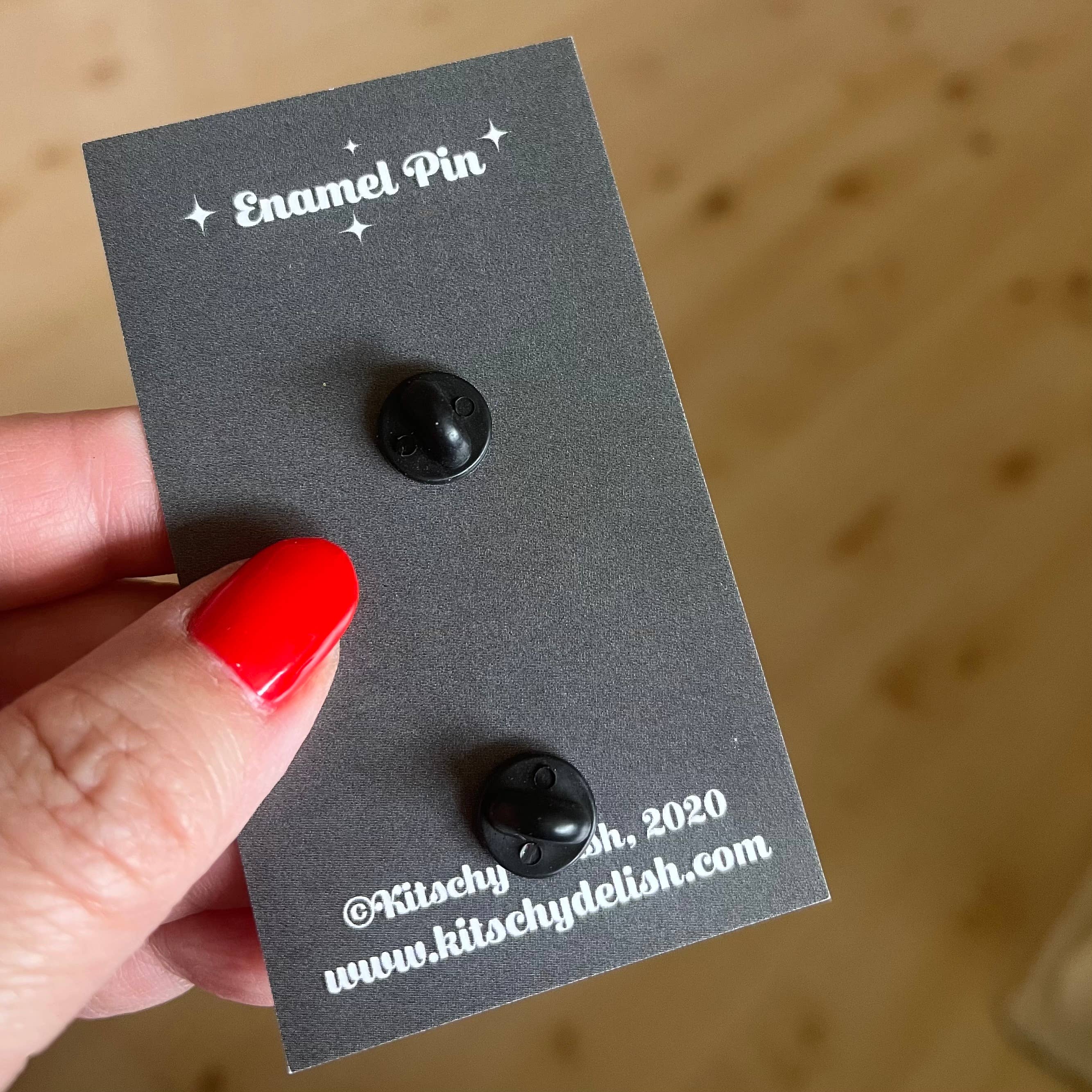Black enamel pin on a gray card held by a hand with red nail polish against a wooden background.