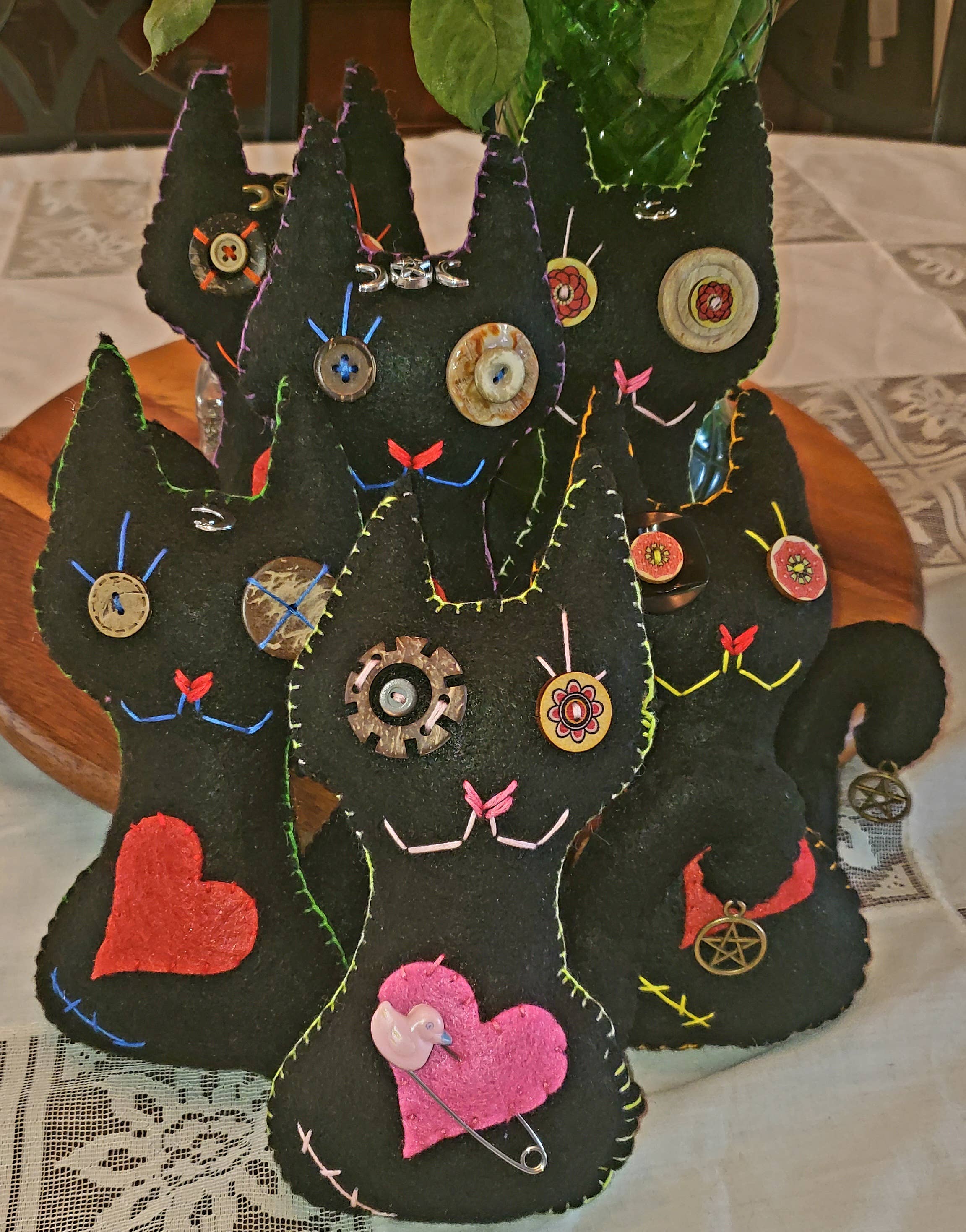 A group of handmade black felt cat figurines decorated with colorful buttons, safety pins, gears, and felt hearts, arranged together on a lace tablecloth.