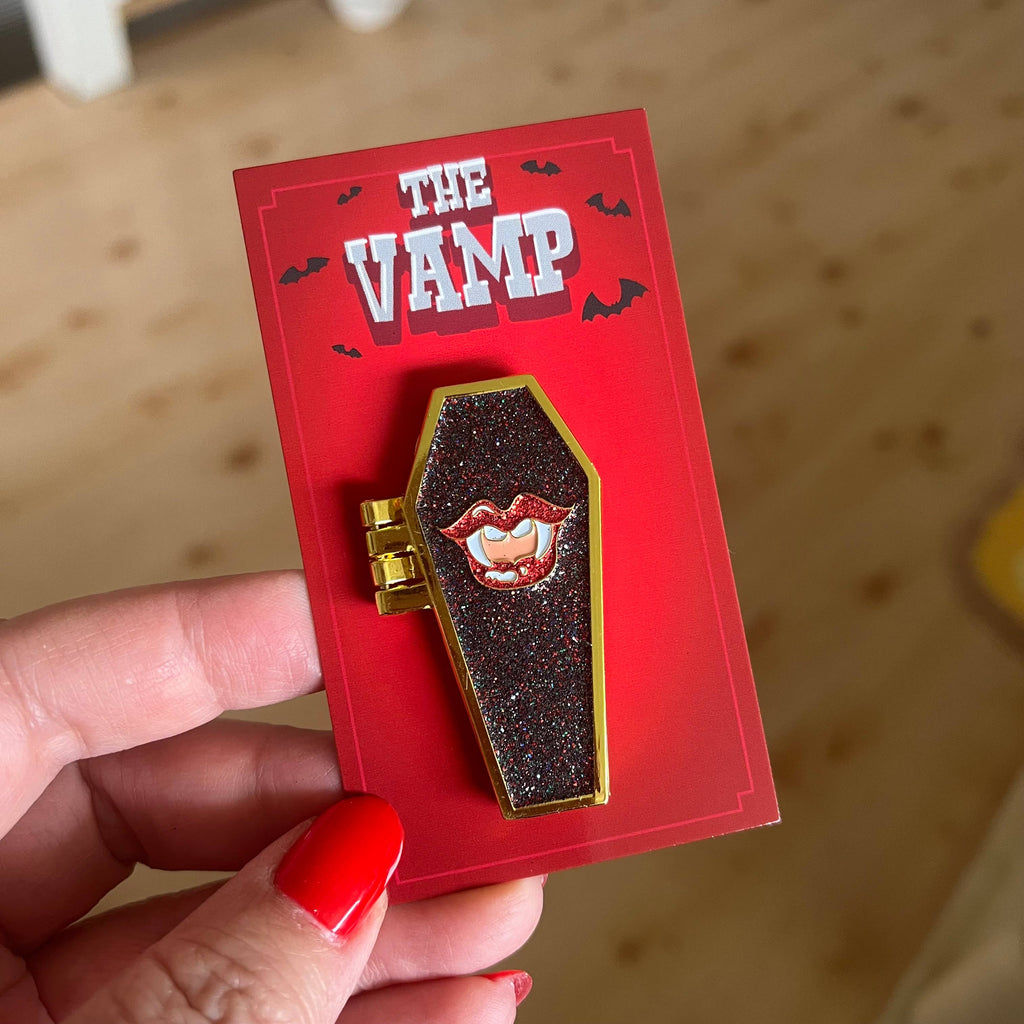 Coffin-shaped pin accessory with glittery texture and red lips design on a red card labeled 'The Vamp'.