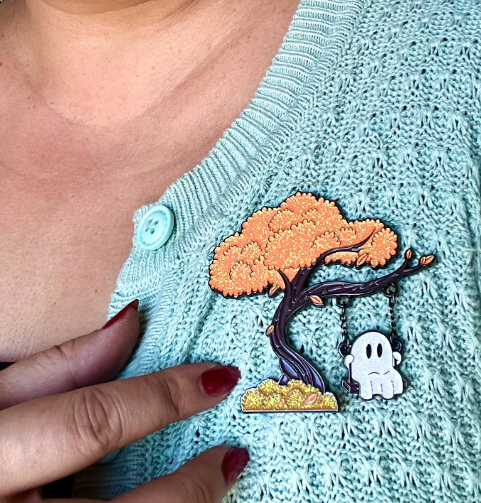 Person wearing a light blue knitted sweater with a tree and ghost enamel pin.